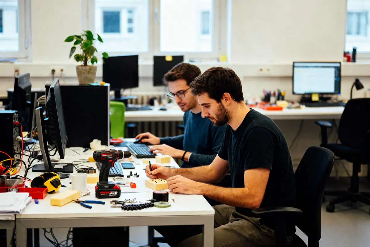 Deux personnes travaillant sur des projets électroniques, entourées d'équipements, dans un bureau avec ordinateurs.